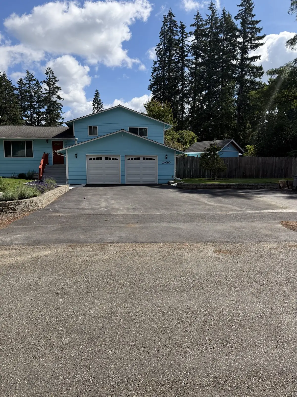 The driveway apron before sealcoating: faded, chalky gray asphalt in front of a blue two-story house.