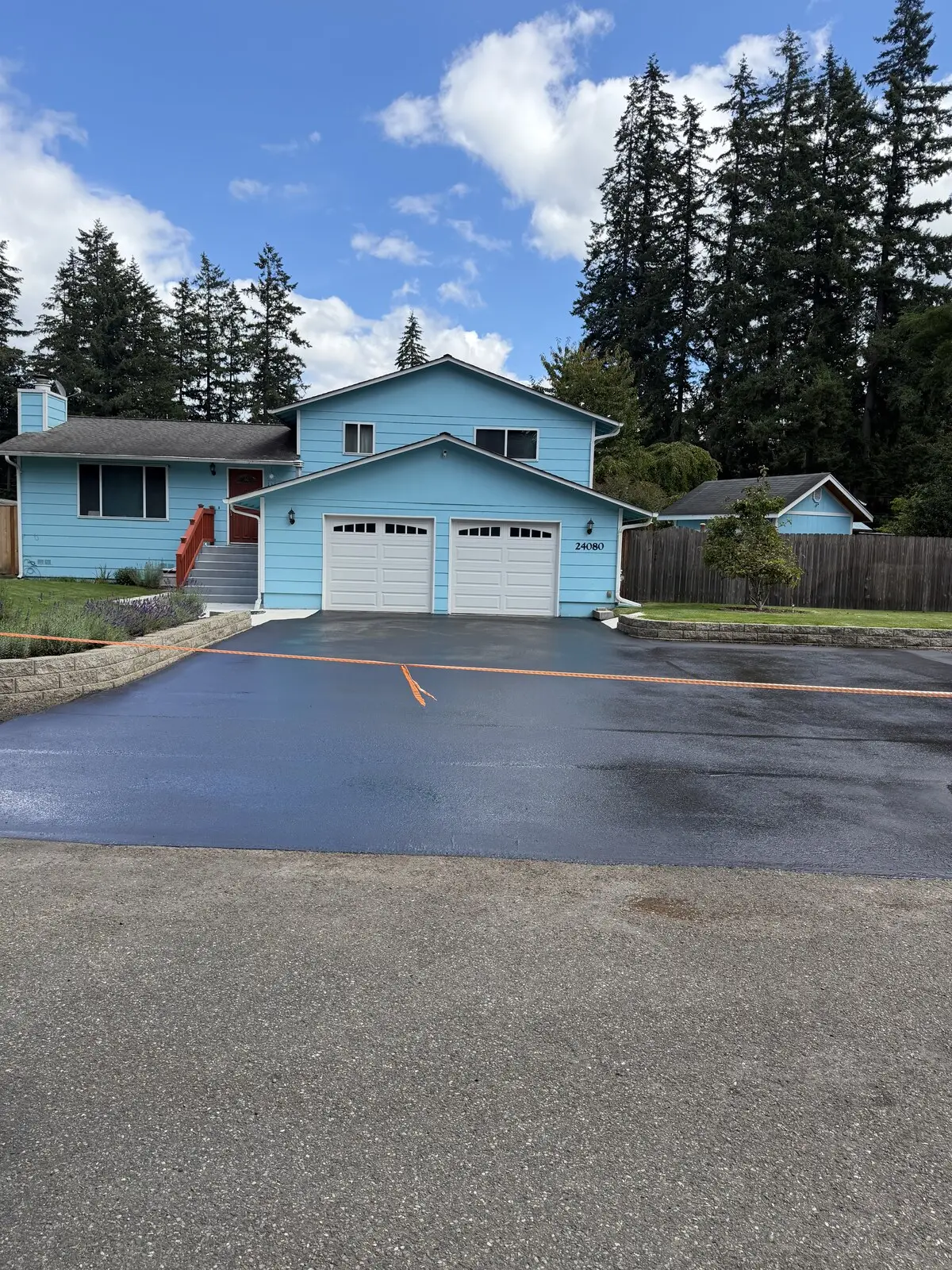 A wide residential driveway apron freshly sealcoated jet black in front of a blue two-story house.