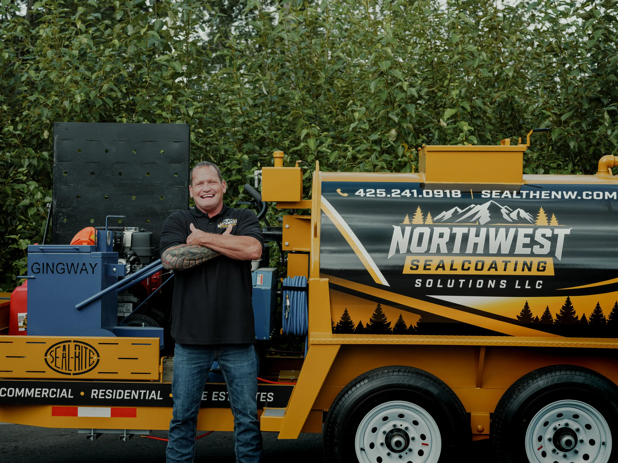Chad standing in front of his fully branded Northwest Sealcoating Solutions trailer, with hose reel and hot crack-fill kettle visible.